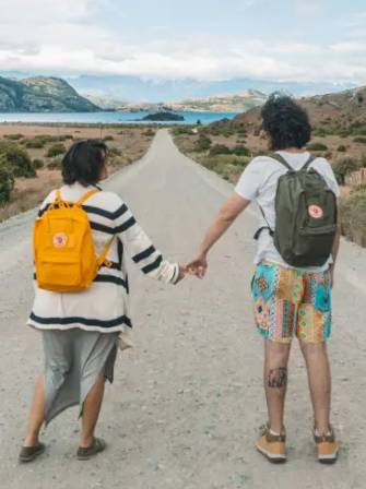 Marcelo e Tainá de mãos dadas com mochilas nas costas em estrada, ao fundo, um lago. Seguro Viagem