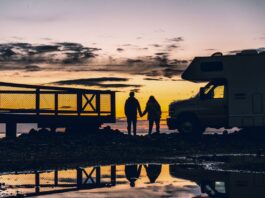 Silhueta de casal de mãos dadas ao pôr do sol no Homer Spit, Alasca, com motorhome ao lado e reflexo na água