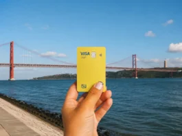Conta Nomad euro: como funciona e vale a pena? Cartão Visa da conta Nomad segurado em frente à ponte 25 de Abril, em Lisboa